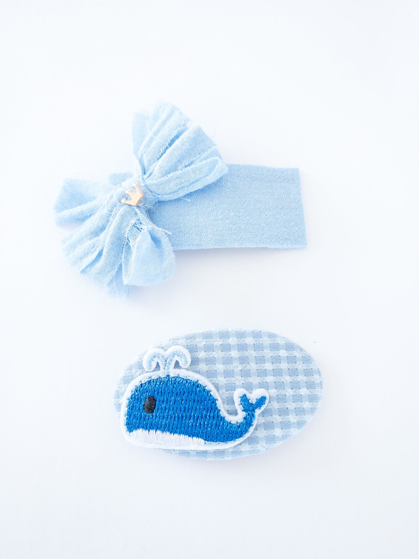 The cutest little blue whale is affixed to a soft blue gingham pattern fabric all wrapped around an oval barrette. The rectangle barrette is wrapped in a solid blue fabric with a fabric bow and a tiny gold crown attached. Both are snap clips, so easy to snap into your hair!