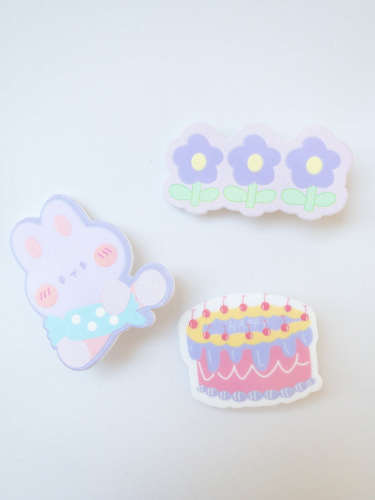 A set of 3 light purple hair clips in the sweetest shapes. A candy holding cartoon bunny, a happy birthday cherry cake and a trio of pretty purple flowers.  Gold alligator clips